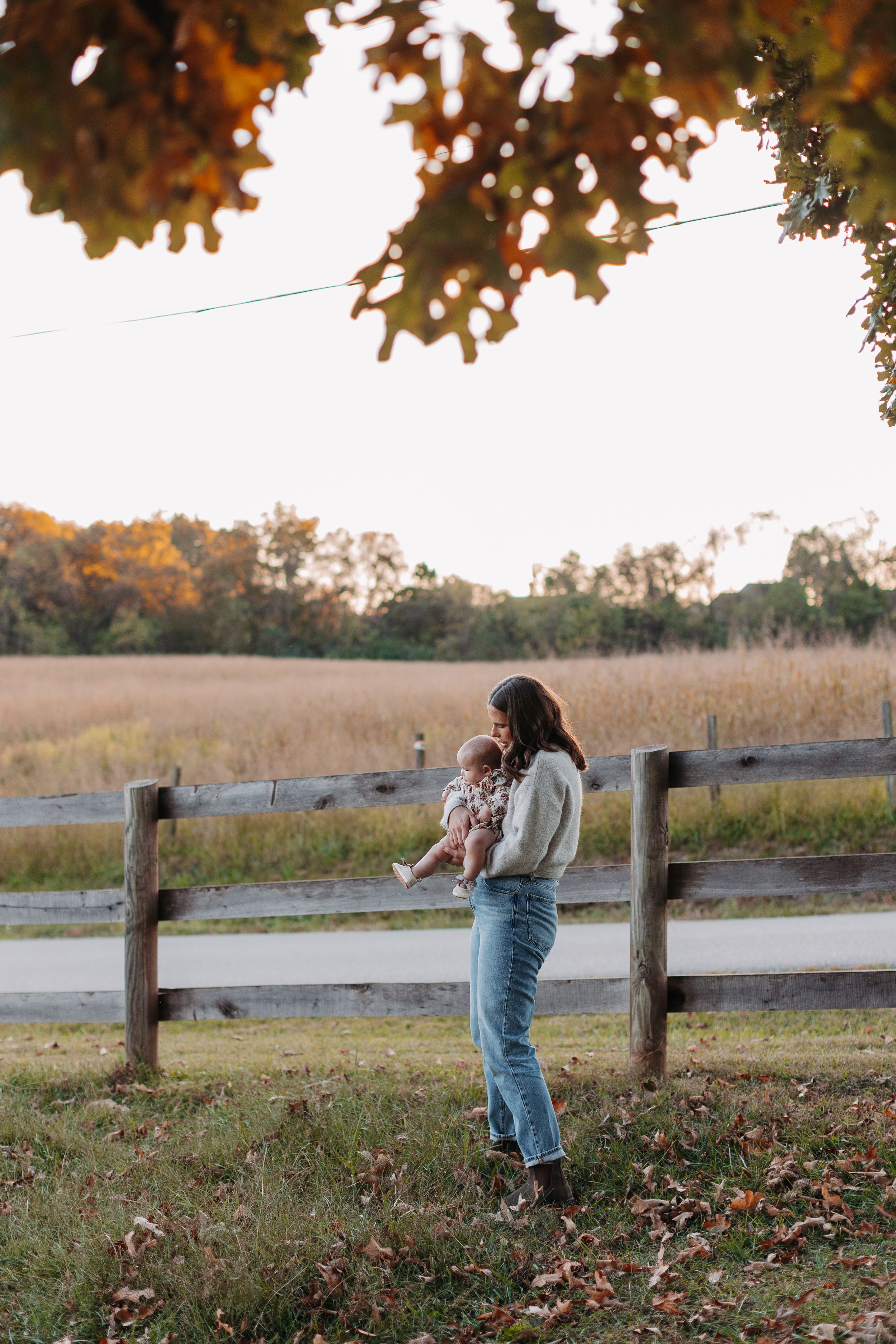 Person in jeans and sweater stands by rustic wooden fence during autumn sunset in a pastoral countryside setting.