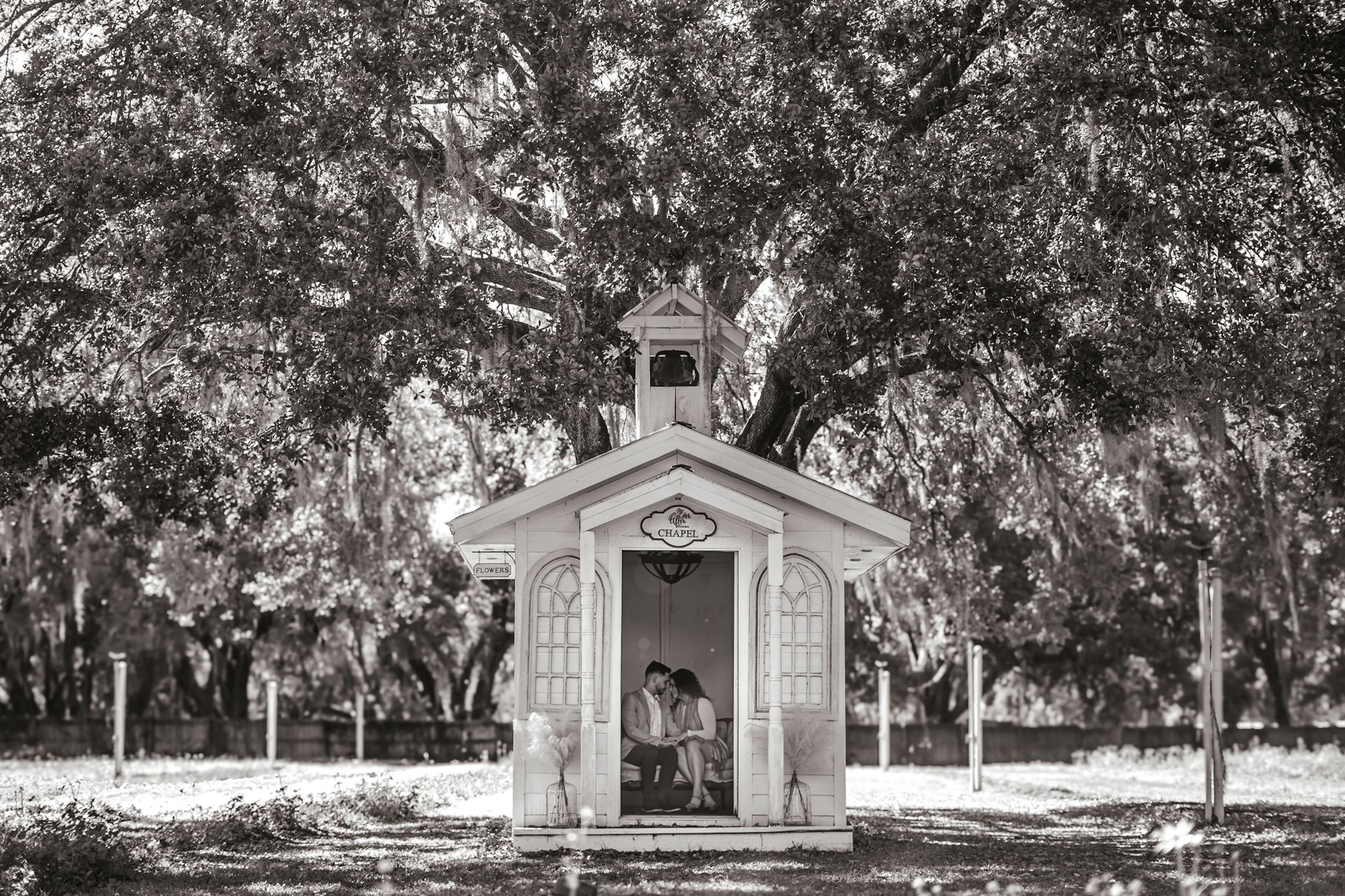 Small white chapel nestled under Spanish moss trees in black and white.