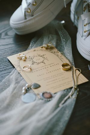 Wedding invitation displayed with white sneakers, jewelry, and delicate tulle fabric on a dark wooden surface.