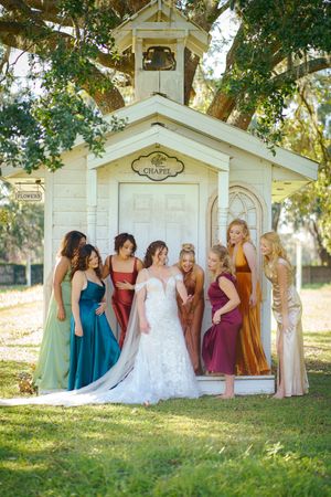 Wedding party poses playfully in front of a white wooden chapel wearing colorful dresses in different shades of blue and pink.