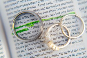 Two wedding bands rest on an open Bible page with text visible through the rings in soft lighting.