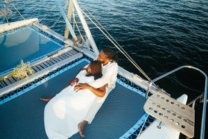 A couple in white outfits relaxing together on the deck of a sailboat on blue ocean waters.