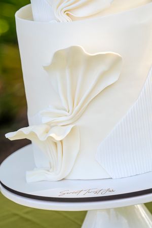 Elegant white wedding cake with delicate pleated fondant draping design detail on a classic cake stand.