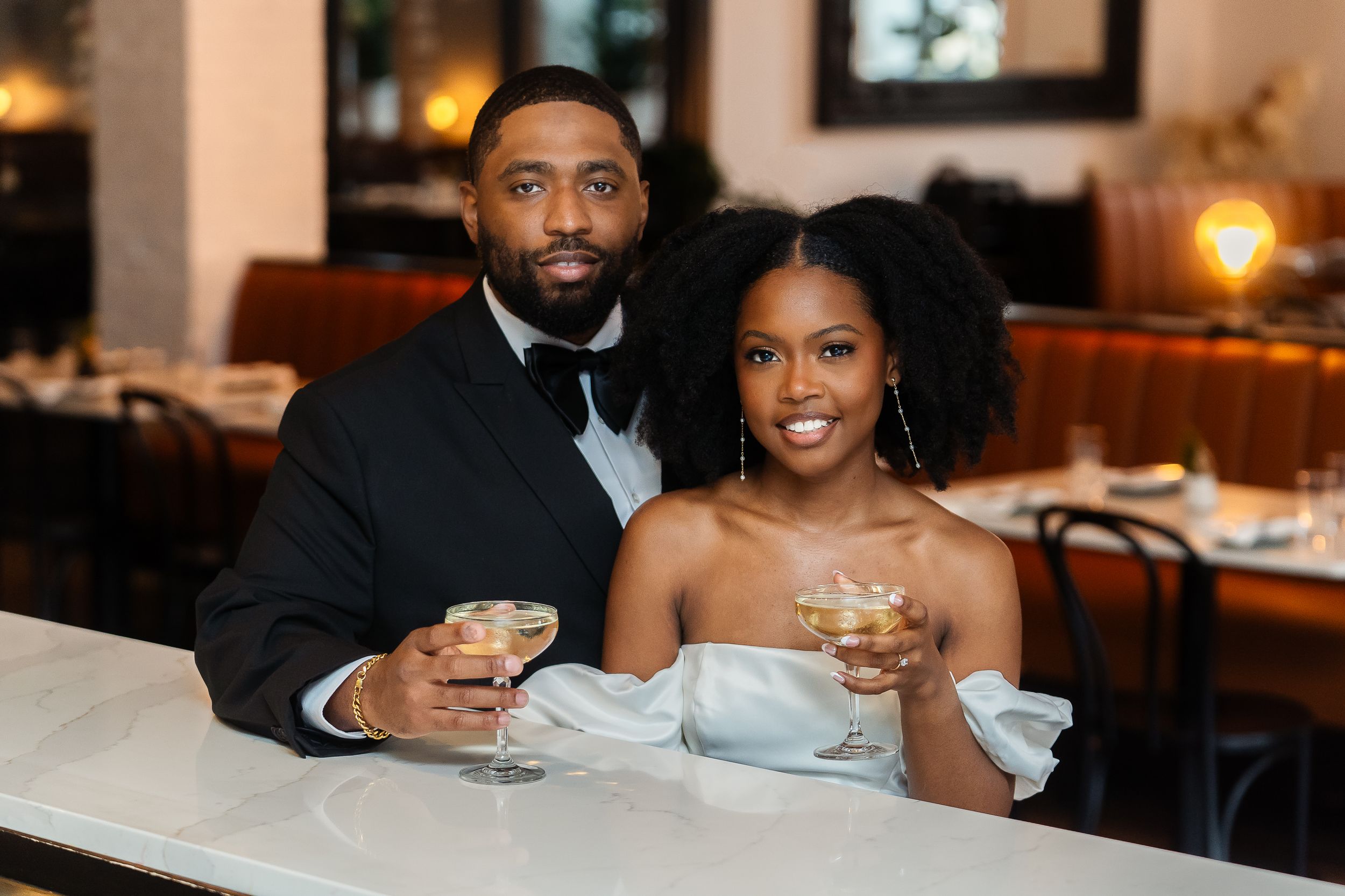 An elegant couple dressed in formal attire shares drinks at a restaurant bar with warm lighting.