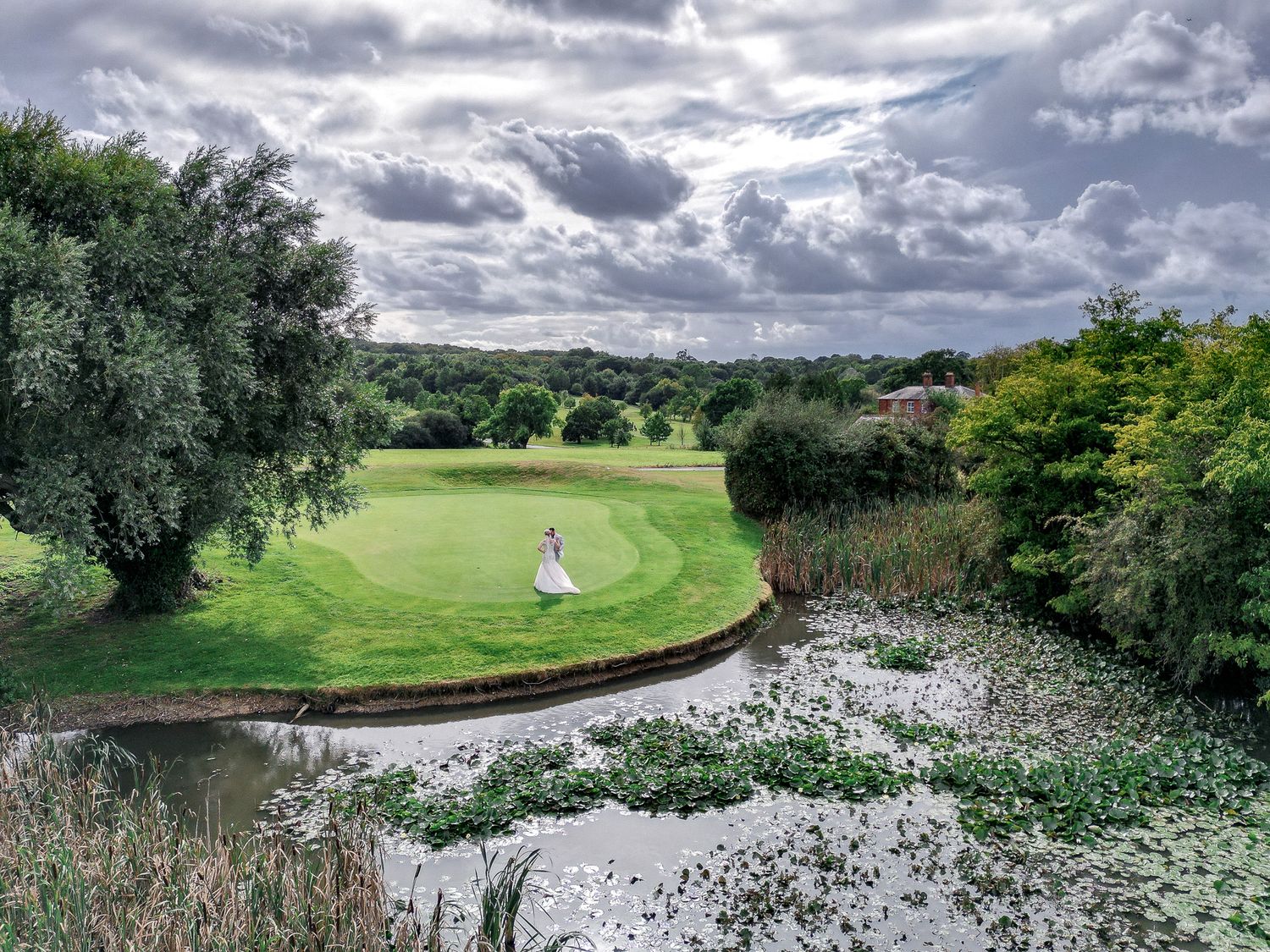 A scenic golf course landscape with lush green fairway curving around a pond under dramatic cloudy skies.