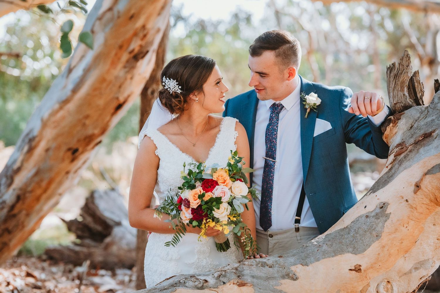 A couple in wedding attire pose together among driftwood at sunset holding a vibrant autumn bouquet.