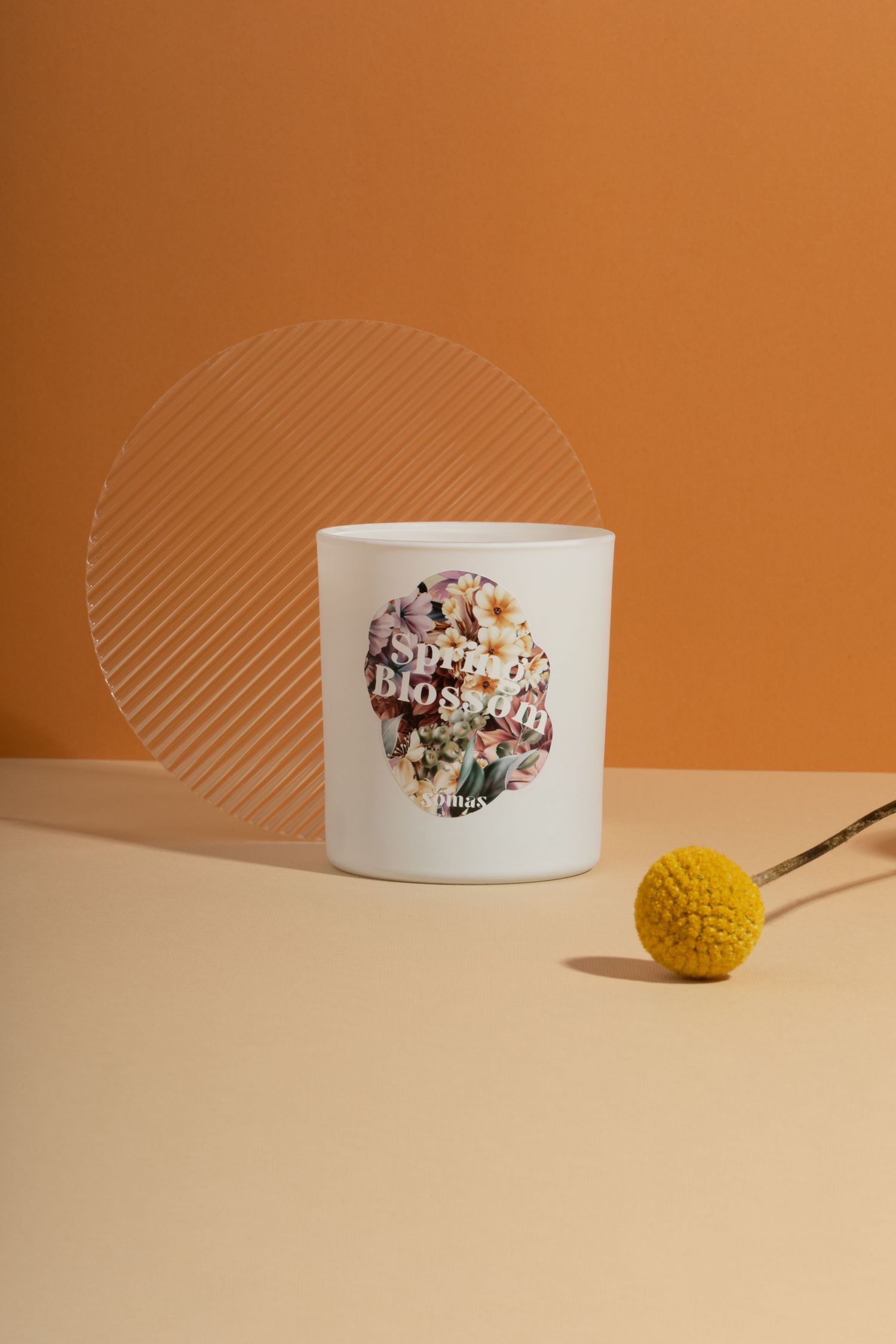 White candle holder with floral design sits beside yellow plant on beige and orange background.
