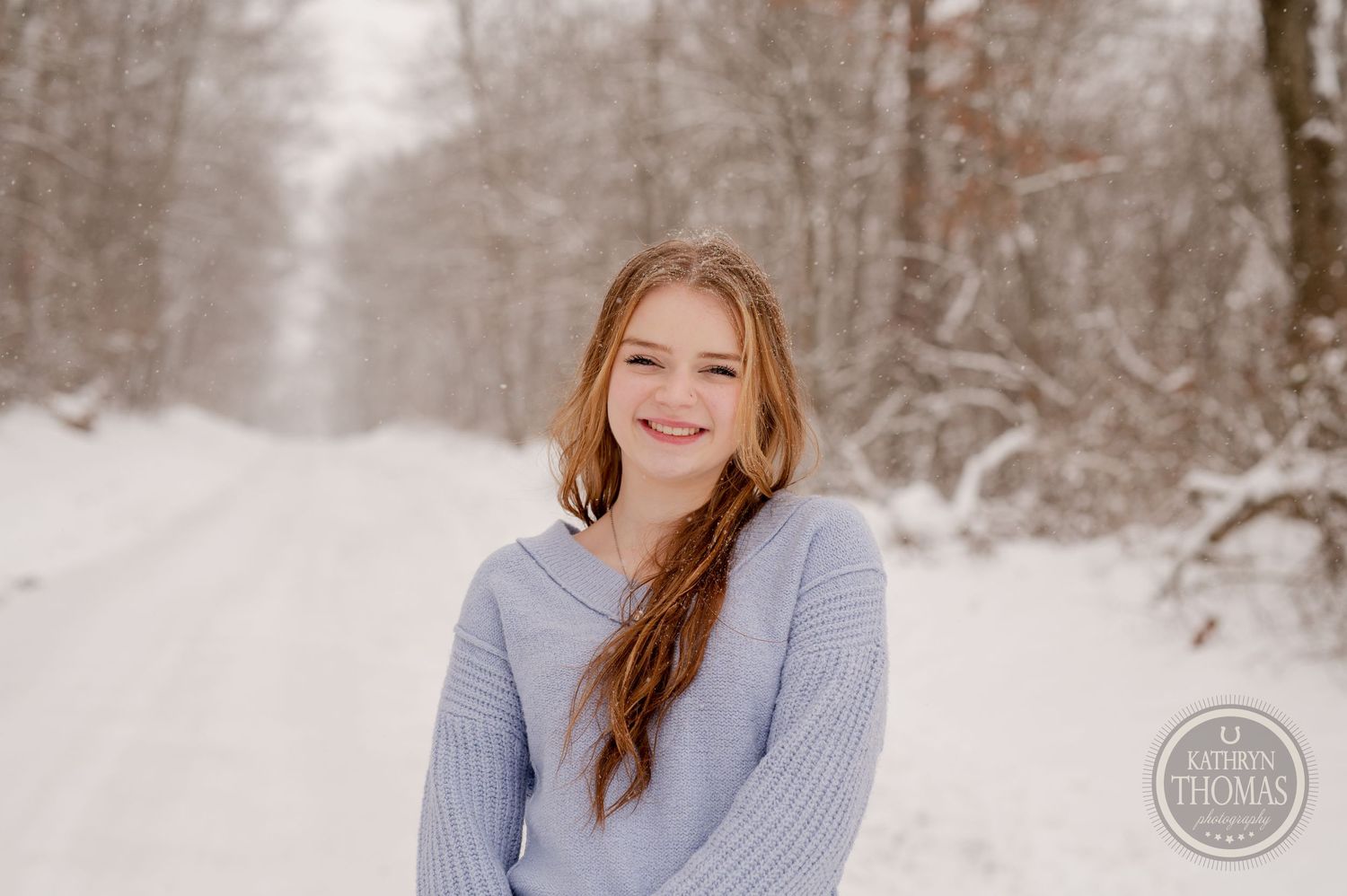 A person in a gray sweater smiling on a snowy winter path surrounded by snow-covered trees.