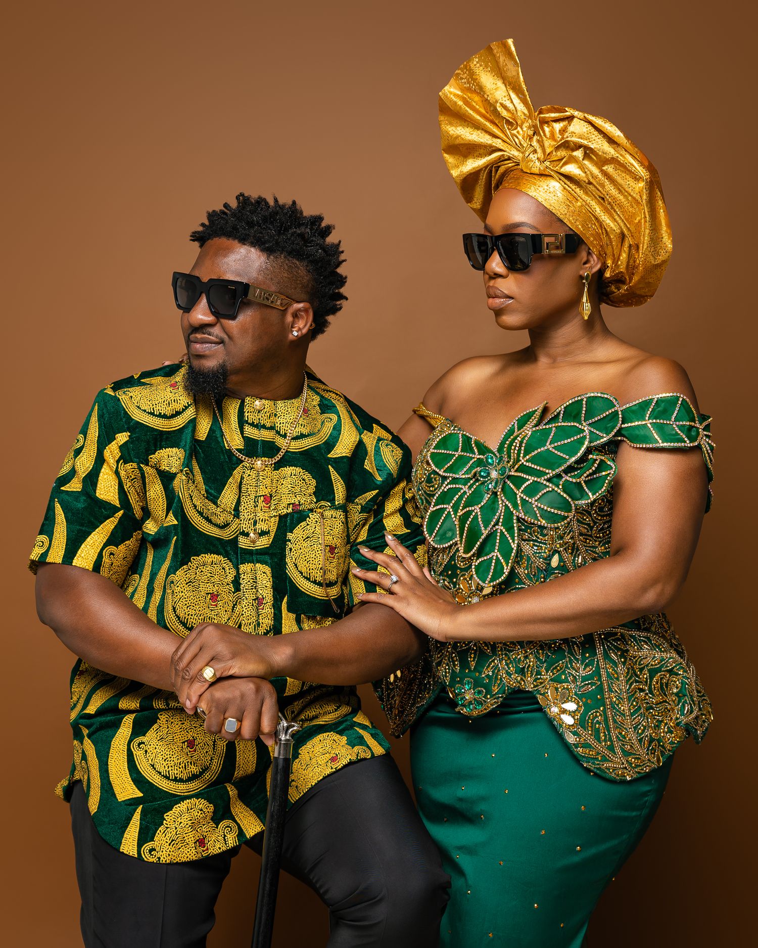 Couple in traditional African attire with bold patterns and headwrap posing against brown background.