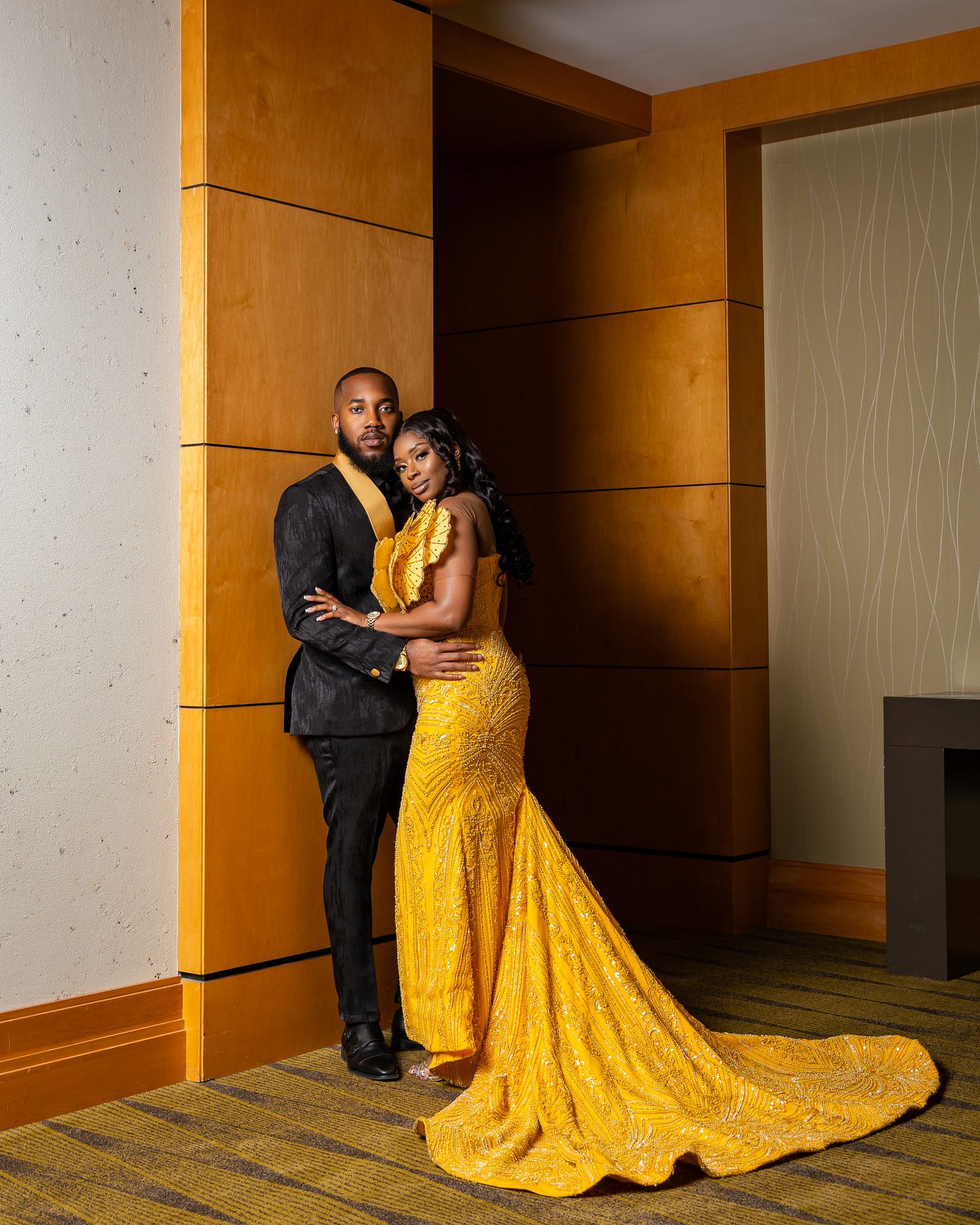 Couple in formal attire posing against wooden paneled wall, woman in yellow gown.