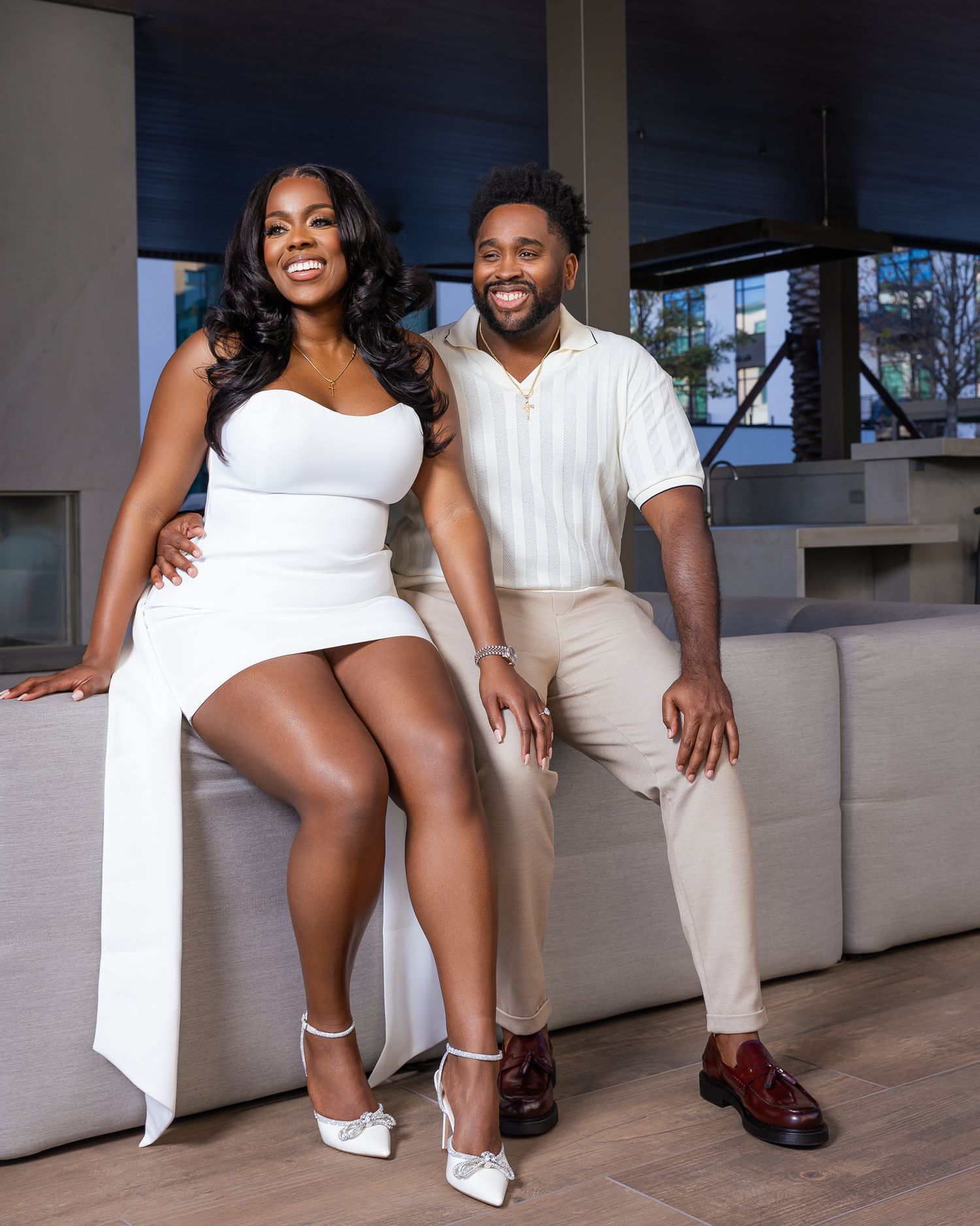 Couple in white outfits posing on a couch in a modern setting.