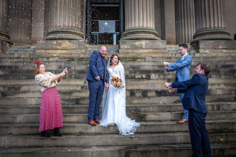 Newlyweds celebrated with confetti shower on stone steps by joyful wedding guests.