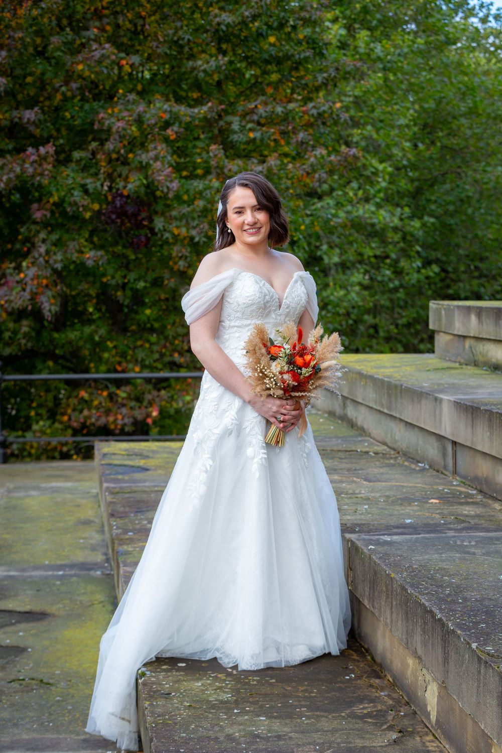 Smiling bride in off-shoulder white tulle gown holding autumn bouquet on stone steps outdoors.