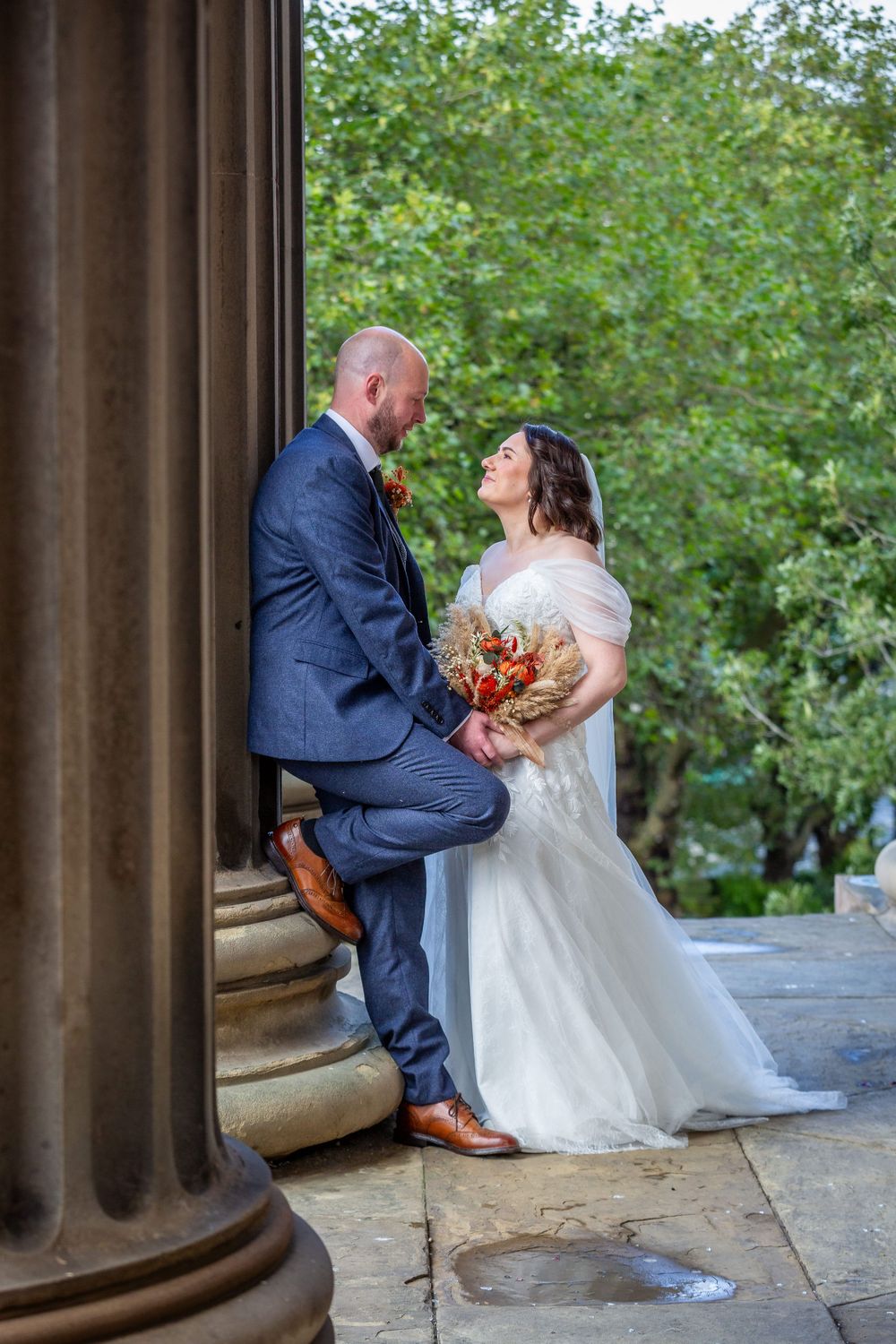 Bride and groom share romantic gaze leaning against classical stone column with autumn bouquet.