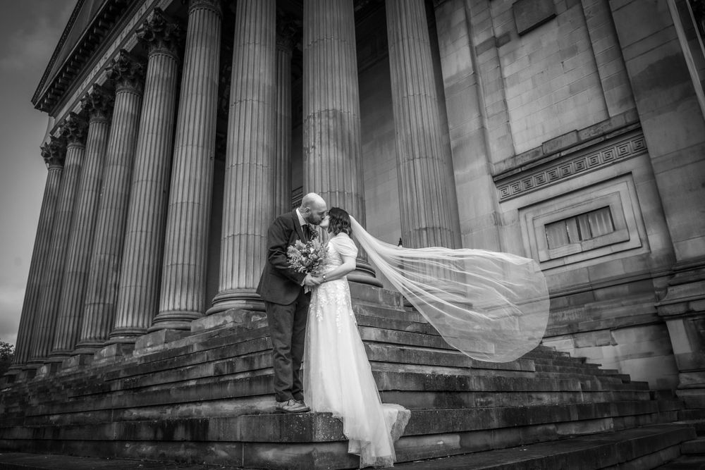 Dramatic black and white photo of newlyweds kissing on steps with veil flowing in wind.