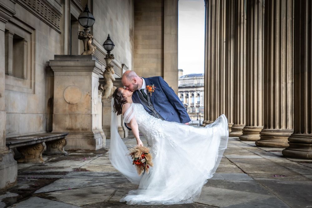 Groom dips bride in elegant dance pose among grand classical columns at wedding venue.