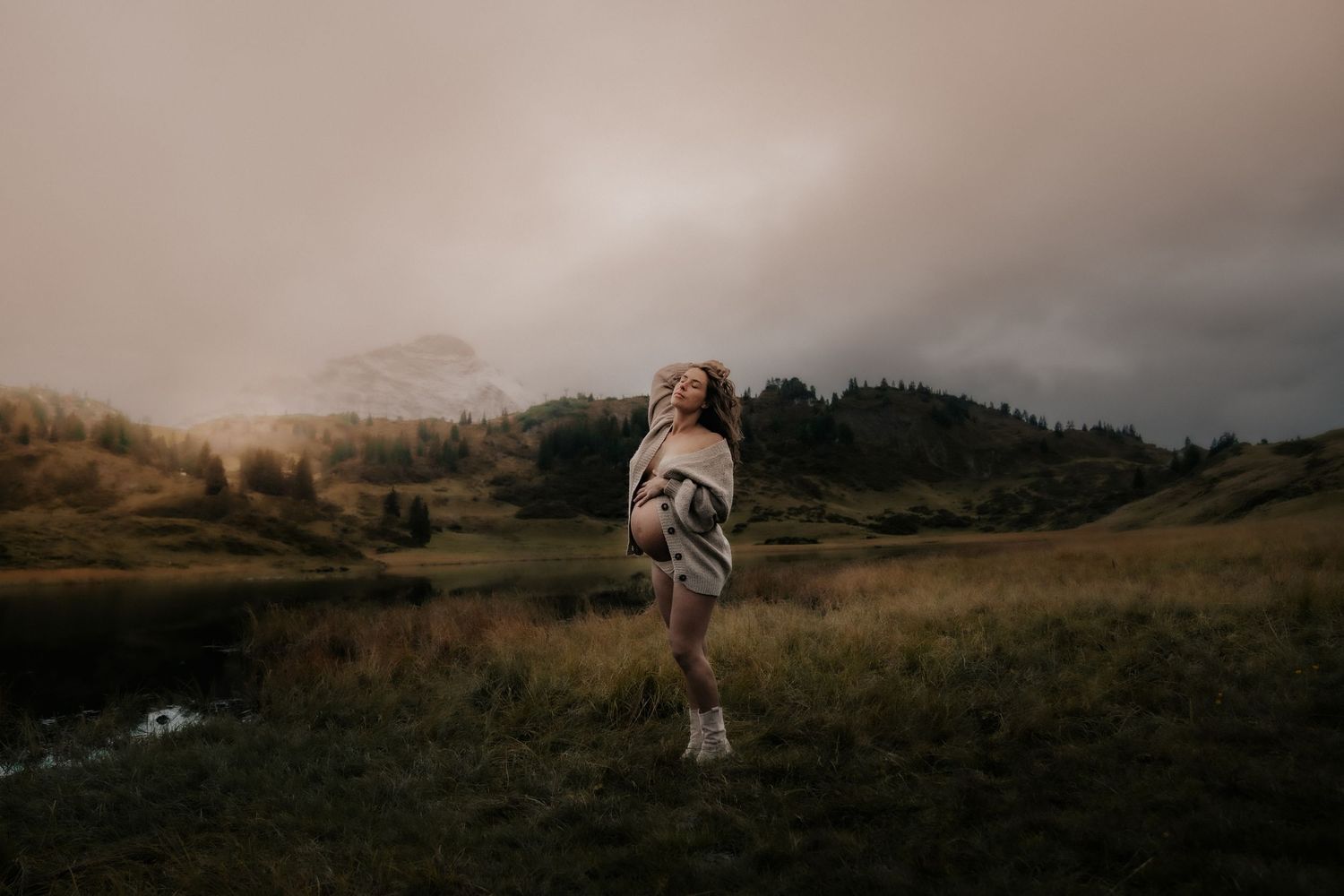A person in a cardigan stands on a grassy hill overlooking a misty mountain landscape at dusk.