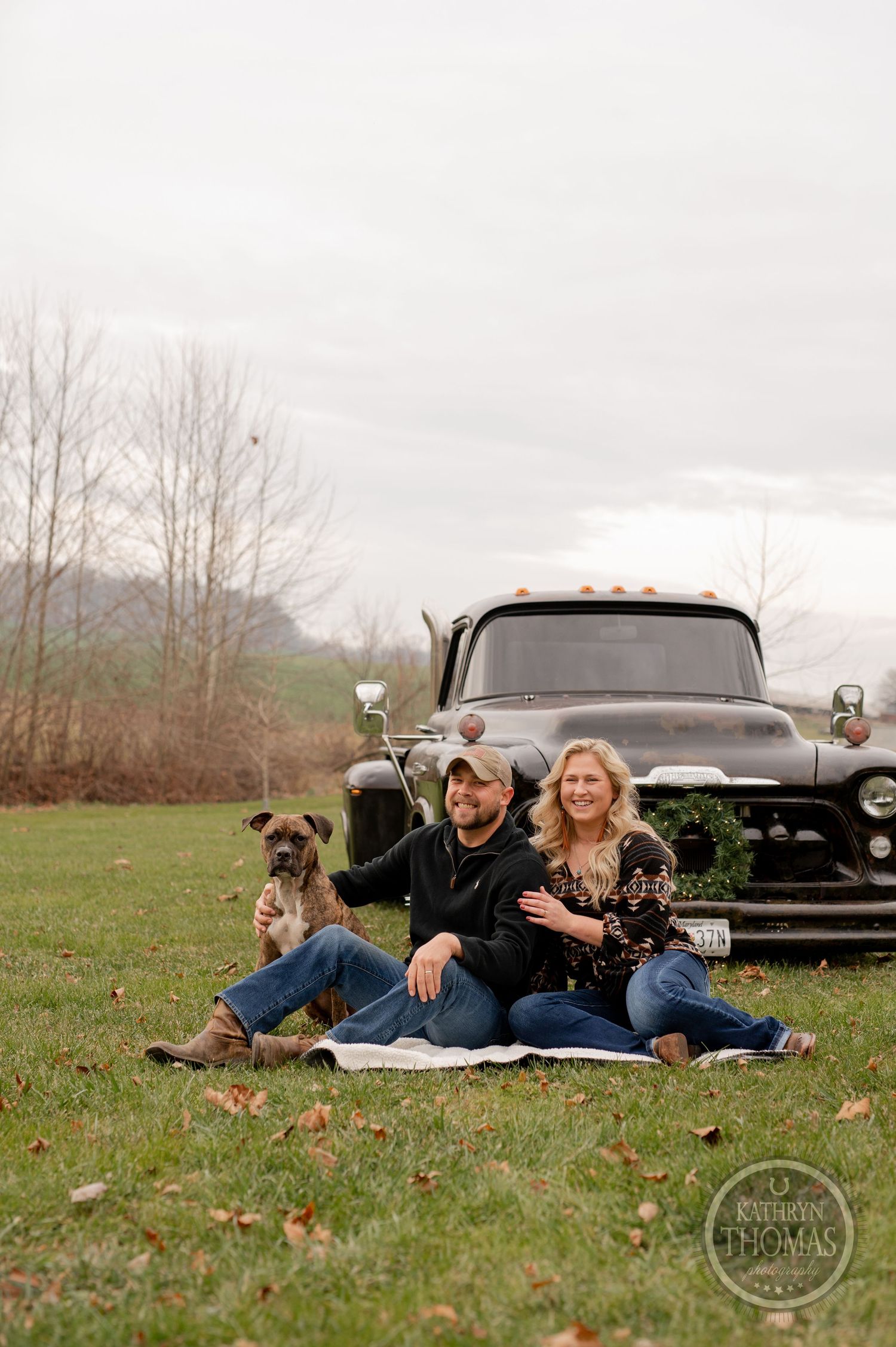 Couple and dog relaxing on picnic blanket by vintage truck in autumn field.