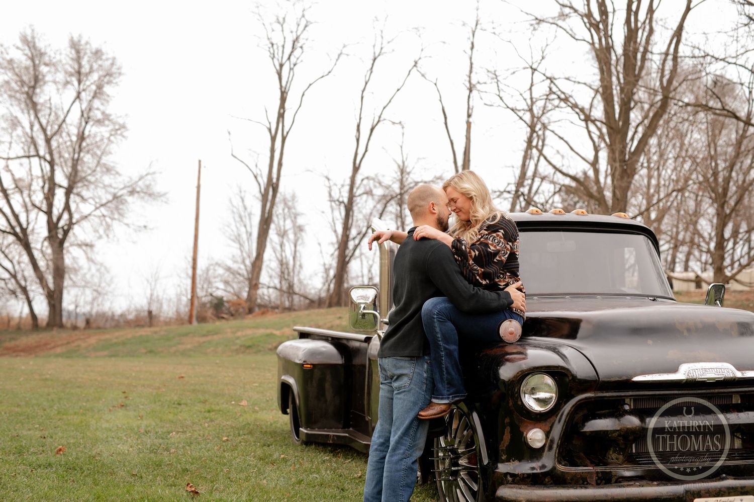 A couple embraces against a classic black pickup truck in a rustic outdoor setting.