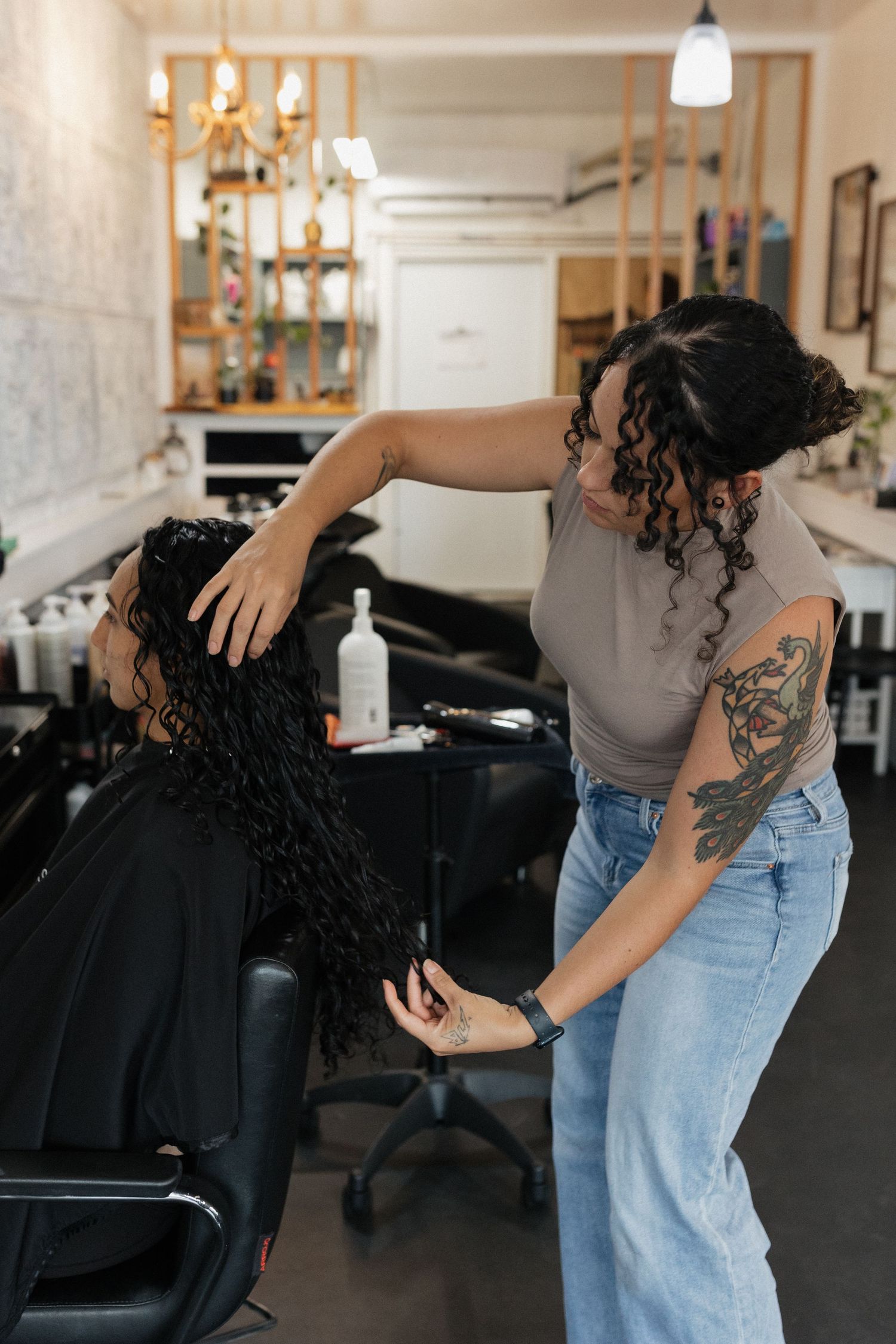 Hairstylist at work in a salon wearing light gray top and jeans with visible arm tattoo styling curly hair.
