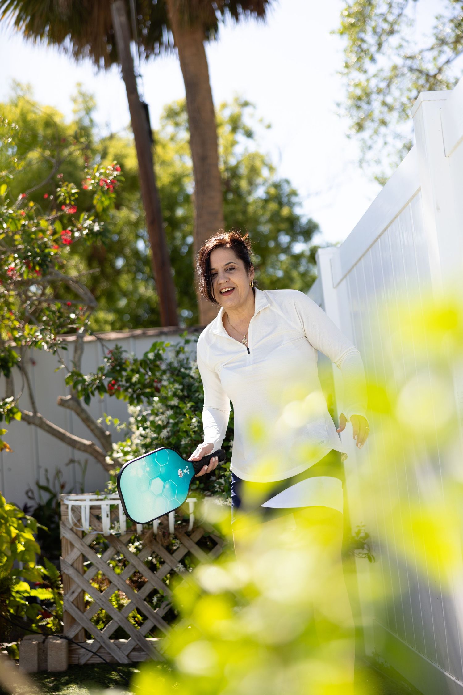 Someone wearing a white collared shirt tends to plants in a sunny tropical garden with a white fence and palm trees.