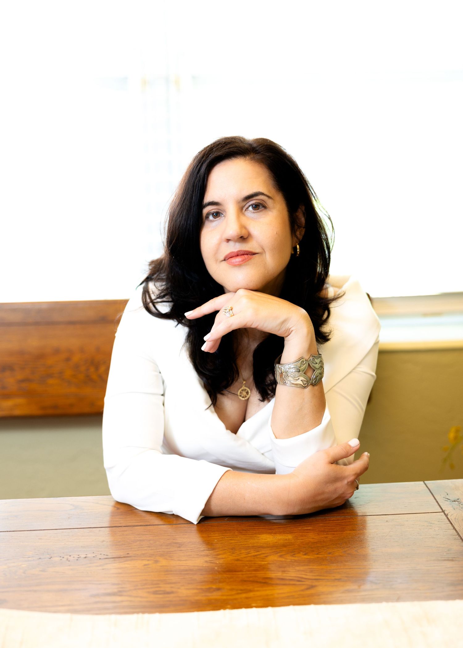 Professional business photo of a person in a white blazer sitting at a wooden table with hands clasped thoughtfully.
