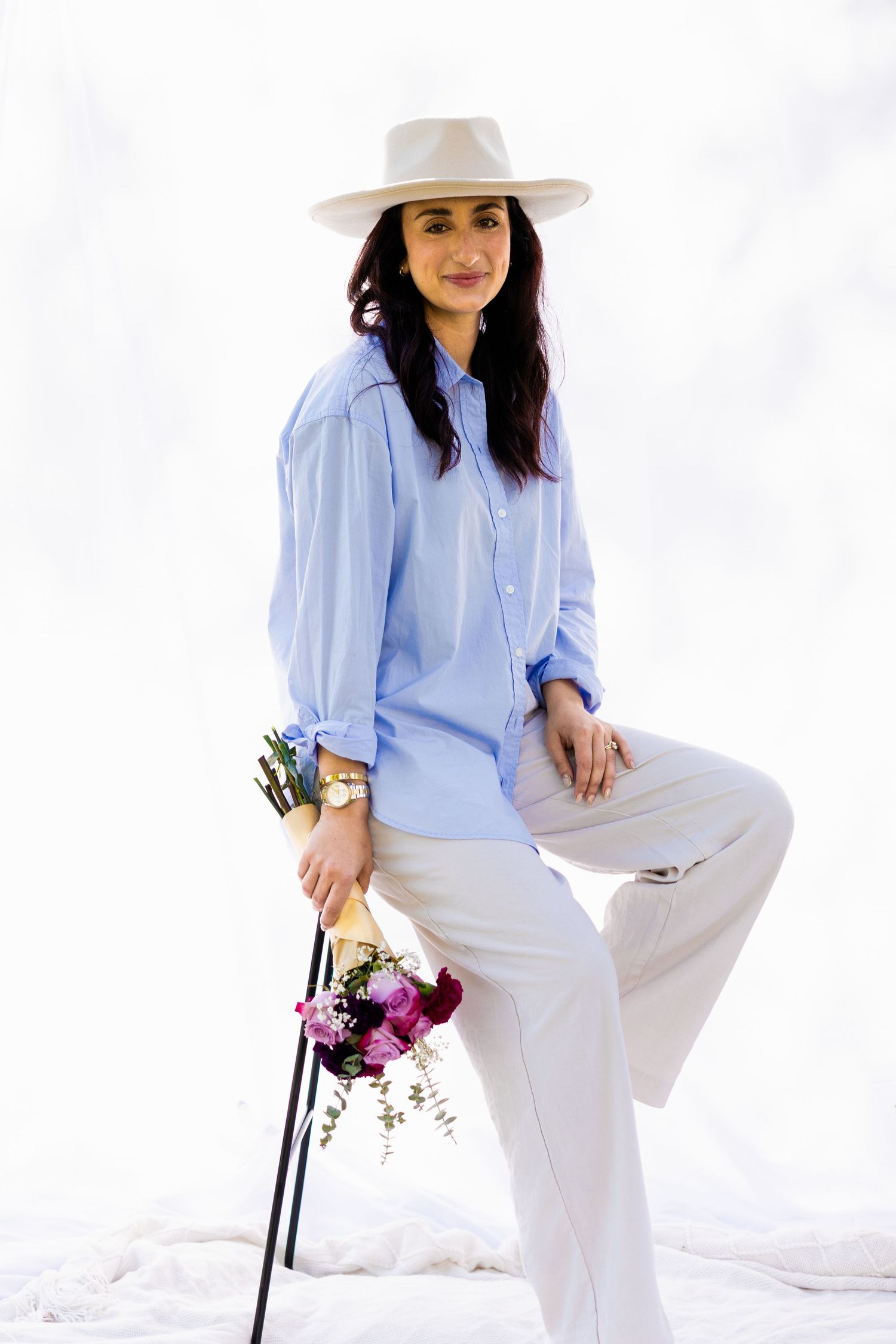 Person in light blue button-down shirt and white pants poses stylishly with flowers while wearing a cream-colored hat.