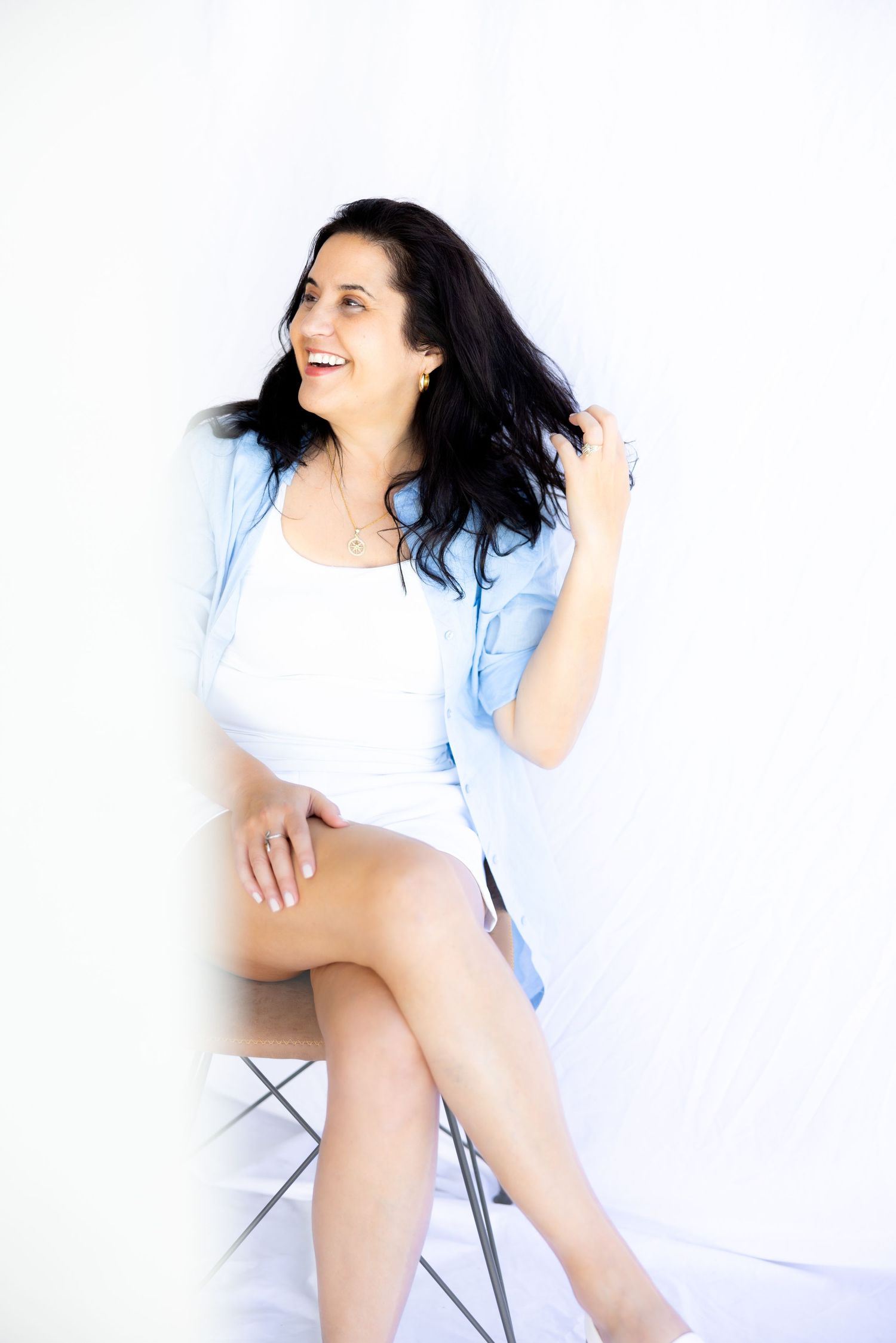 Person wearing white top and light blue shirt poses casually while seated on a chair against white background.