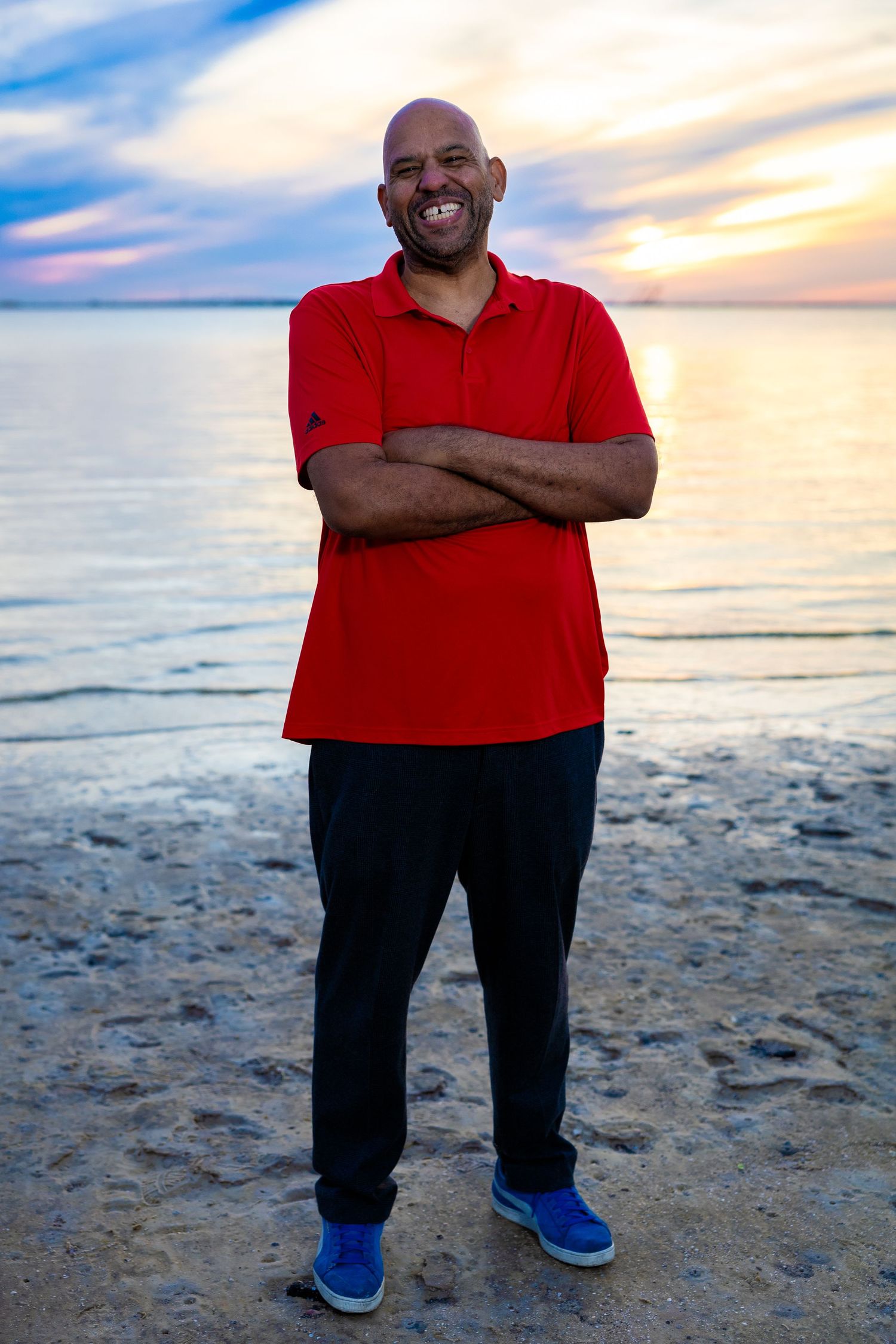 Person in red polo shirt smiling with arms crossed at sunset on a beach.