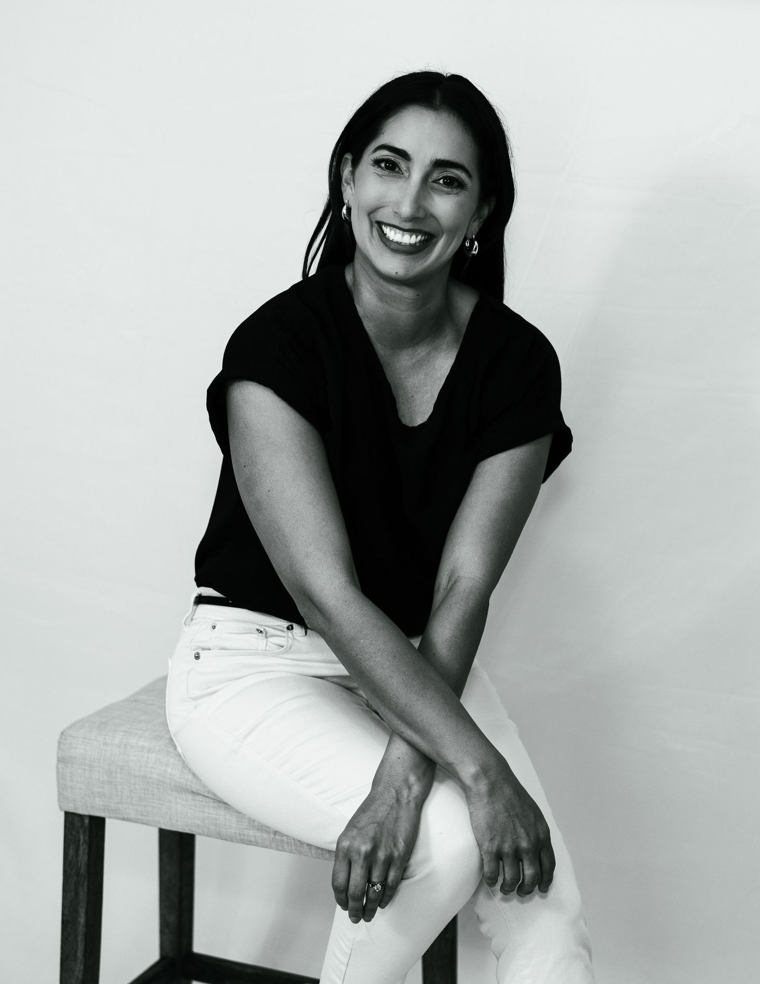 Professional portrait of a person in black top and white pants sitting casually on a chair against white backdrop.