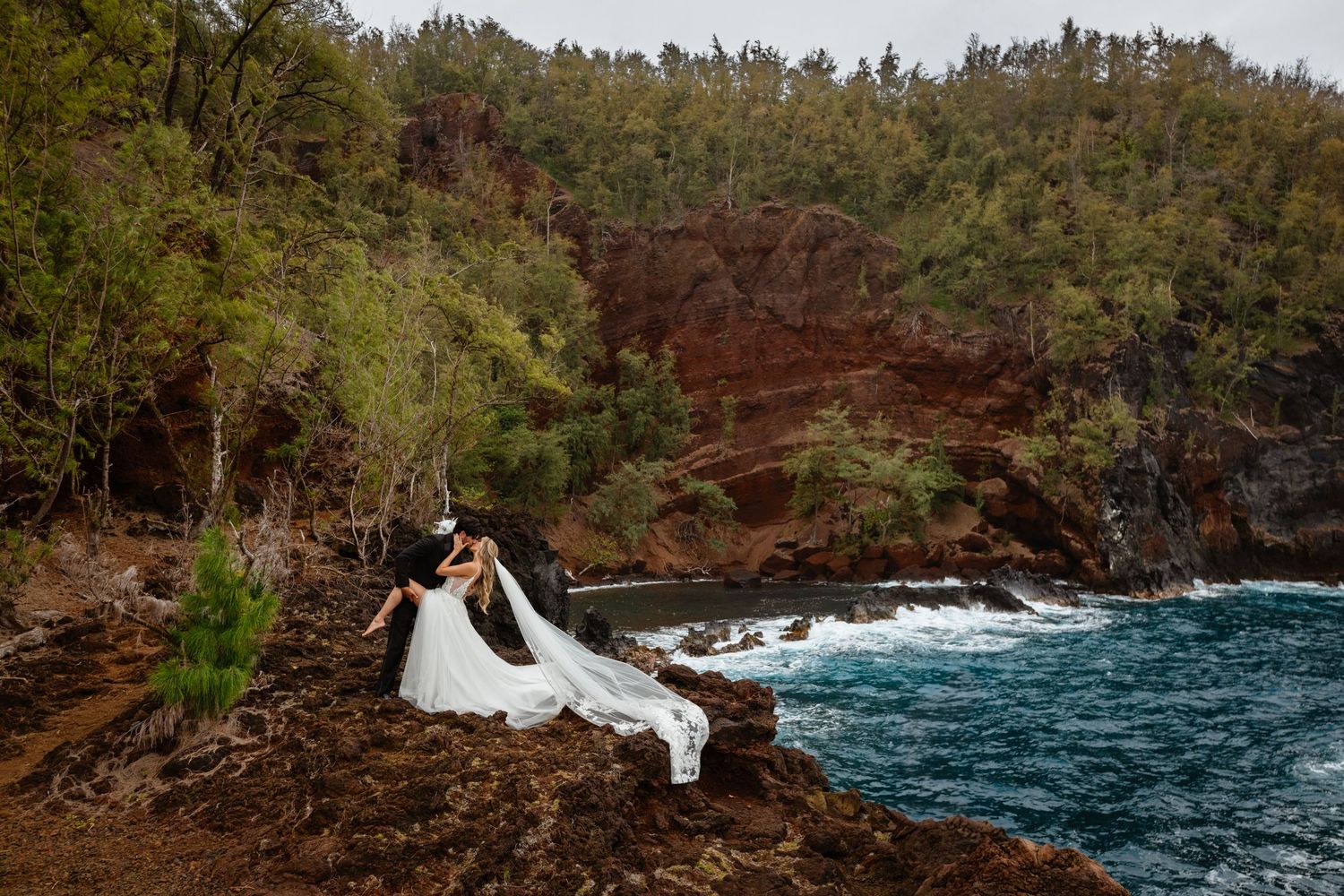 Bride and groom kissing on red volcanic rocks above vivid blue coastal waters surrounded by lush green forest.