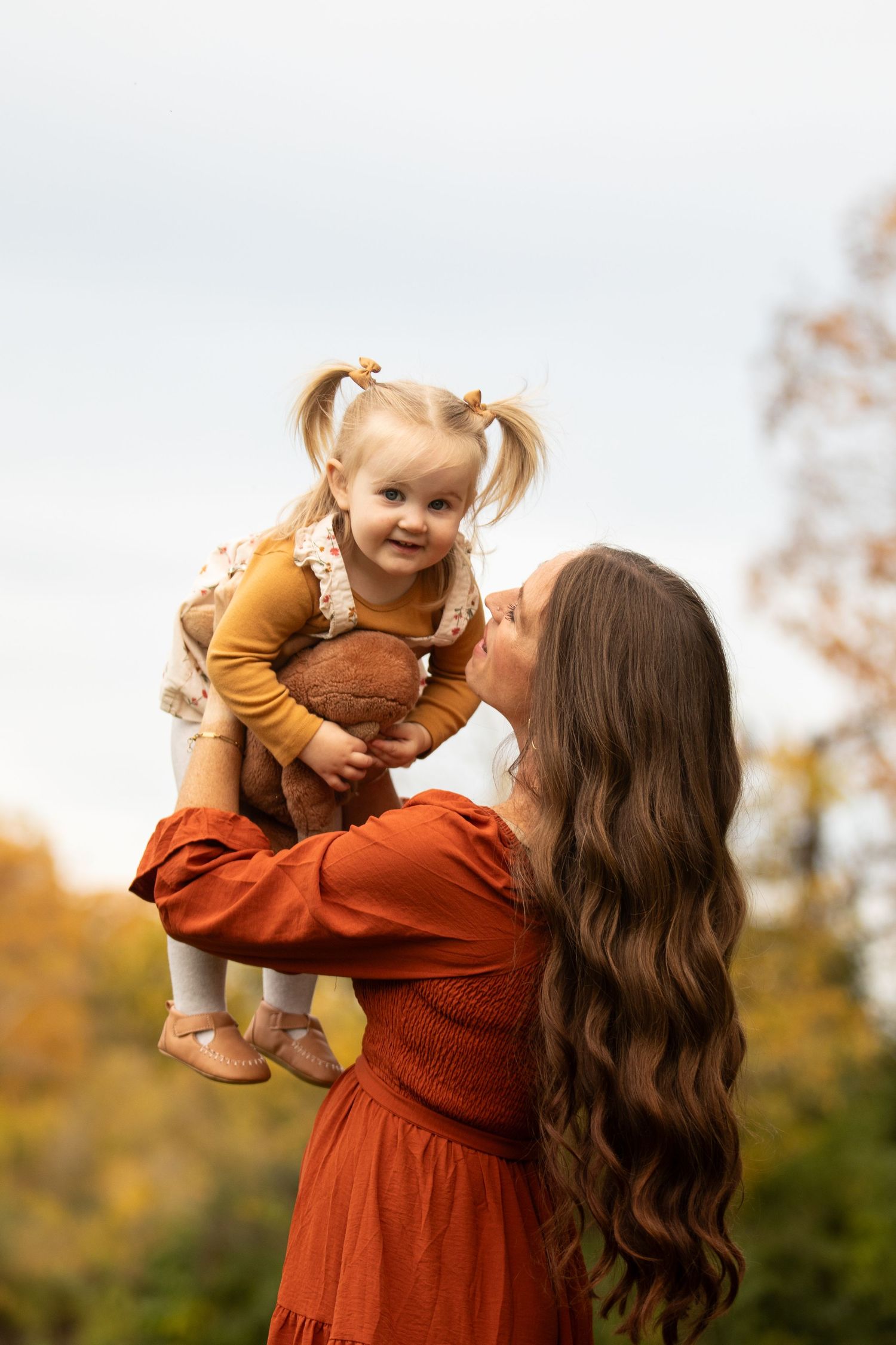 Mother in rust-colored dress lifts and plays with baby wearing brown sweater during autumn outdoor photoshoot.