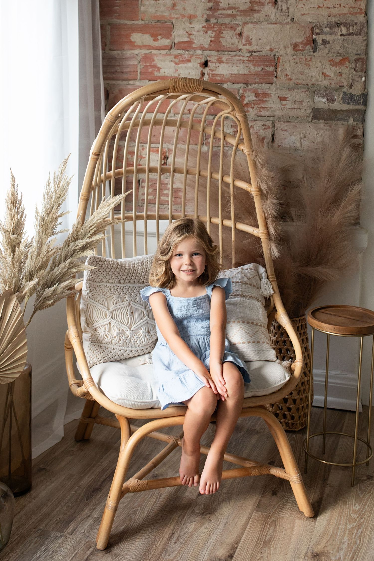 A rattan papasan chair with cushions sits against an exposed brick wall with dried pampas grass decor.