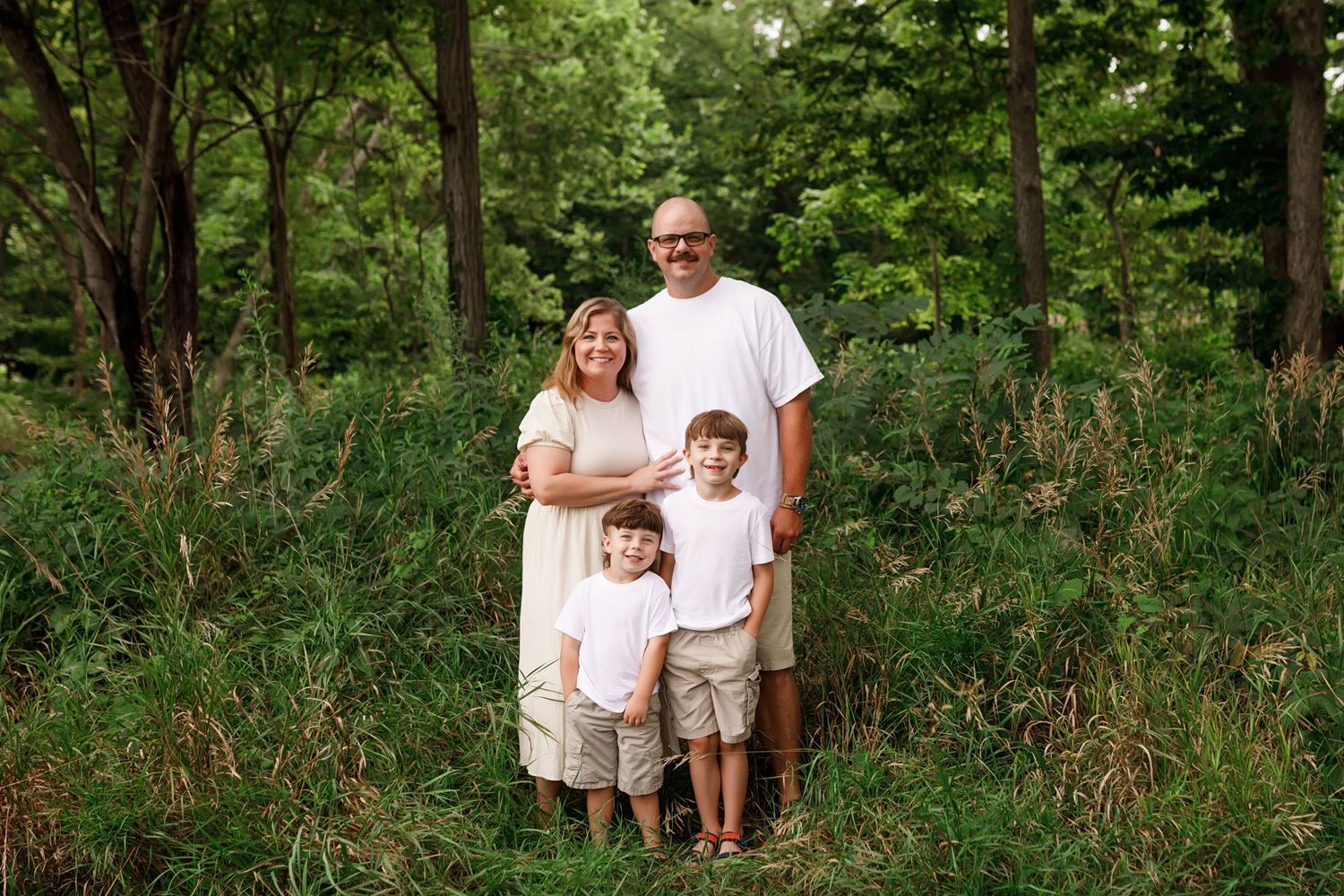 A family in white clothing stands together near lush green trees during an outdoor photo session.