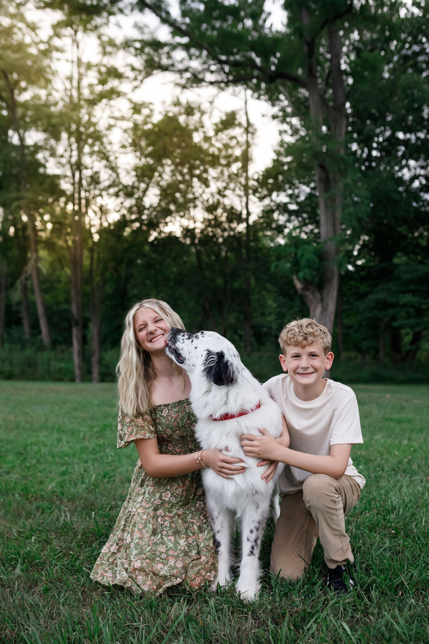 Two siblings playing and smiling with their fluffy white Old English Sheepdog on a grassy lawn at sunset.