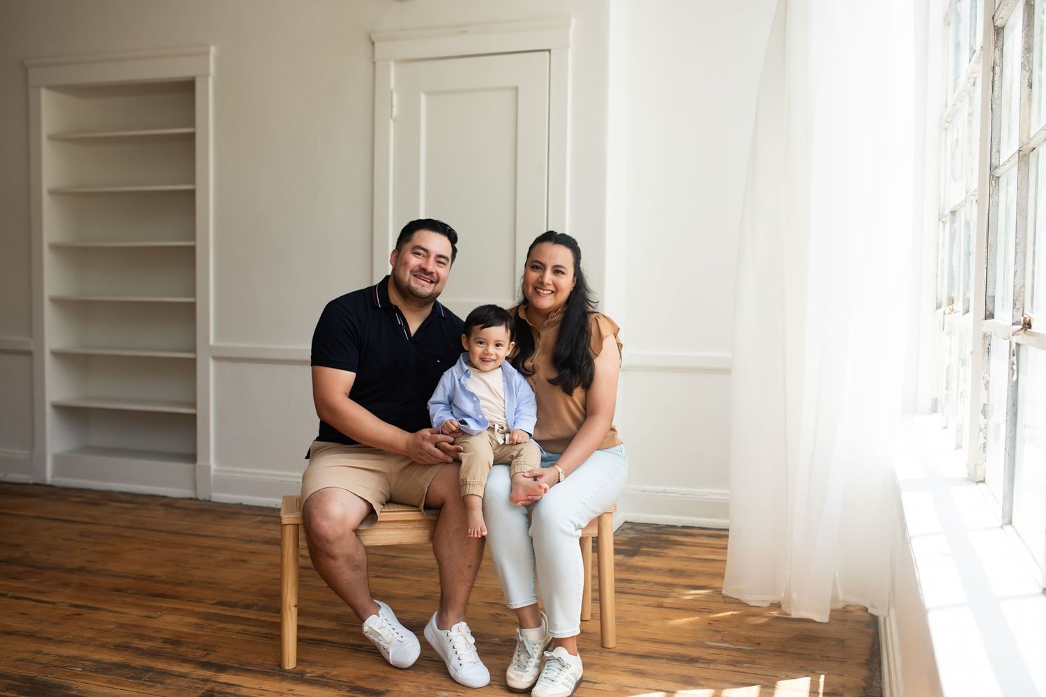 A family sits together on a wooden chair in a bright minimalist room with hardwood floors and built-in shelving.