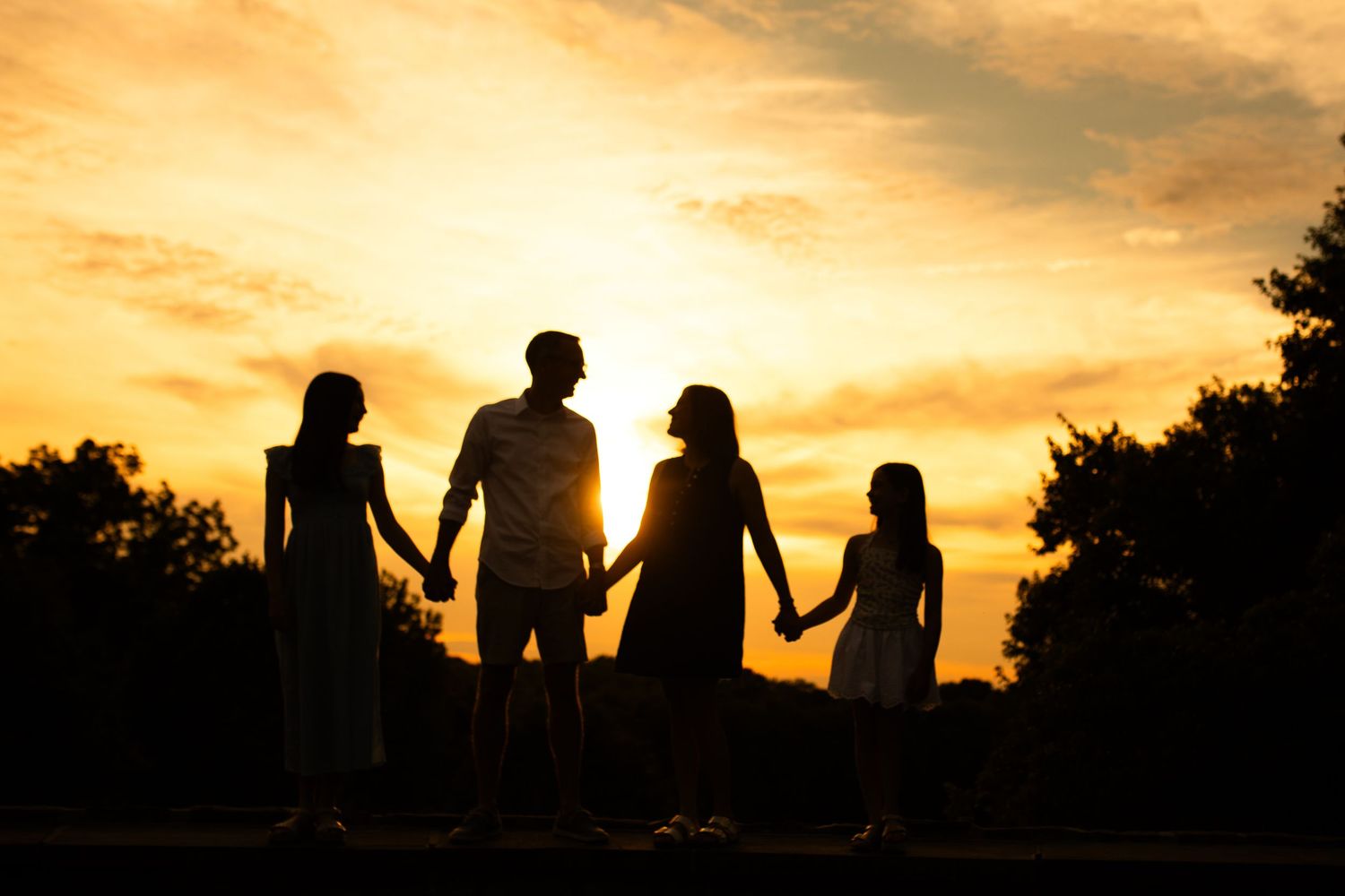 Silhouette of a family of four holding hands against a vibrant golden sunset sky with trees in background.