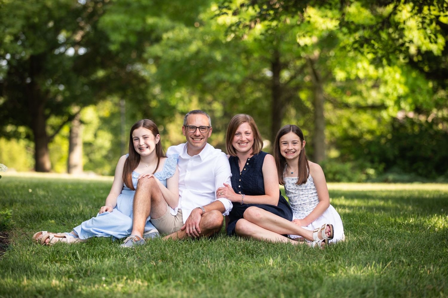 Happy family of four sitting on green grass in a sunny park, smiling together outdoors.