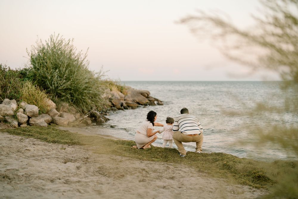 Two figures sit on a wooden bench at sunset overlooking a rocky coastline with shrubs and ocean waves in view.