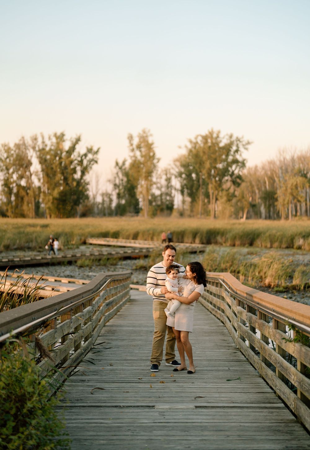 A couple stands together on a wooden boardwalk surrounded by tall grass and trees during golden hour.