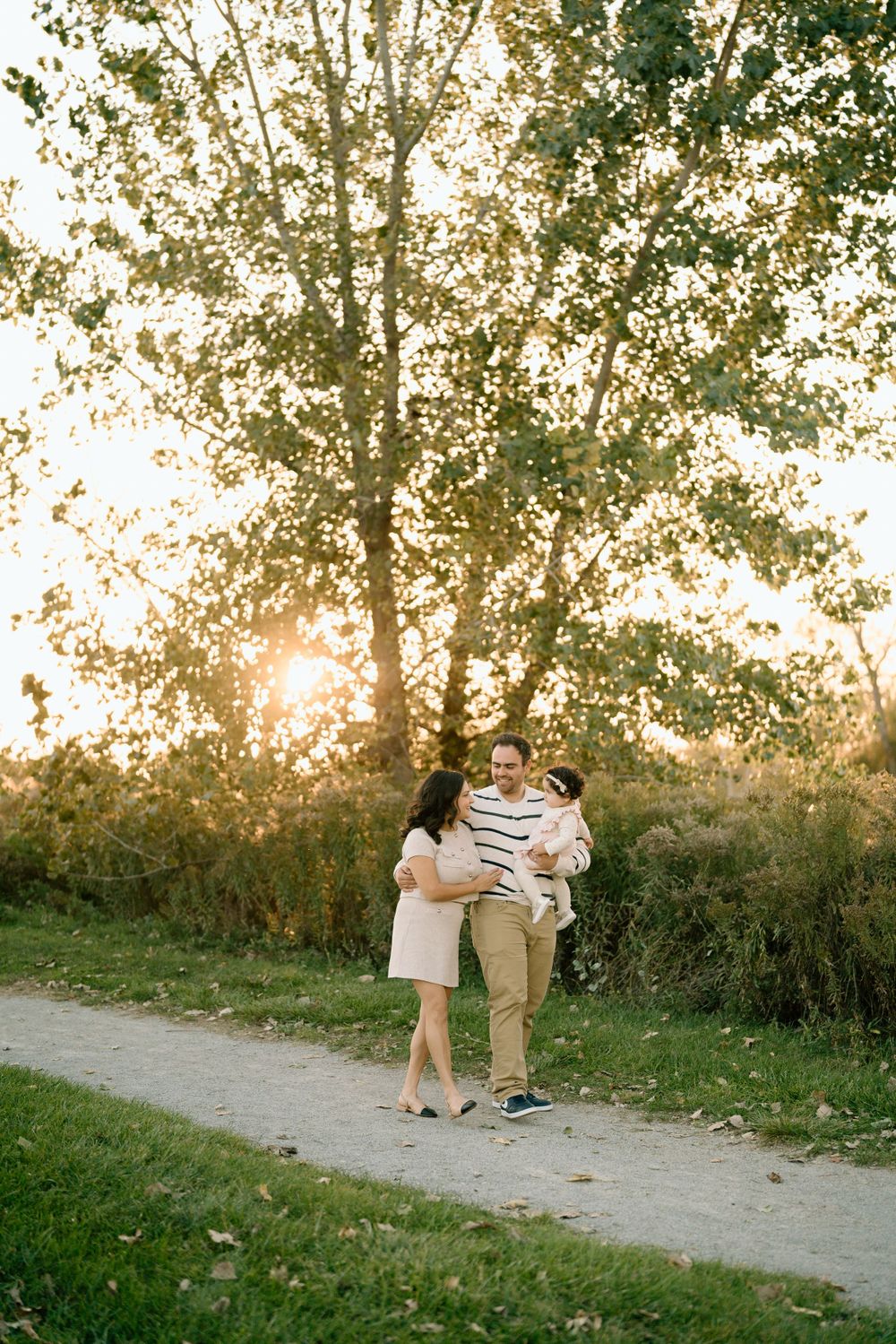 A family stands together on a path at sunset with backlit trees creating a golden glow in the background.