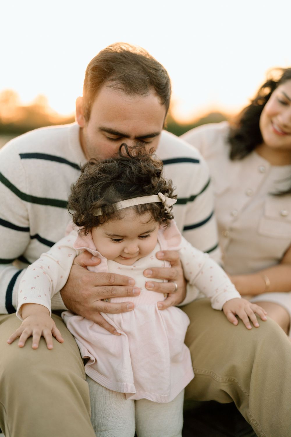 A tender family moment captured at sunset with a toddler wearing a pink shirt and small floral headpiece.