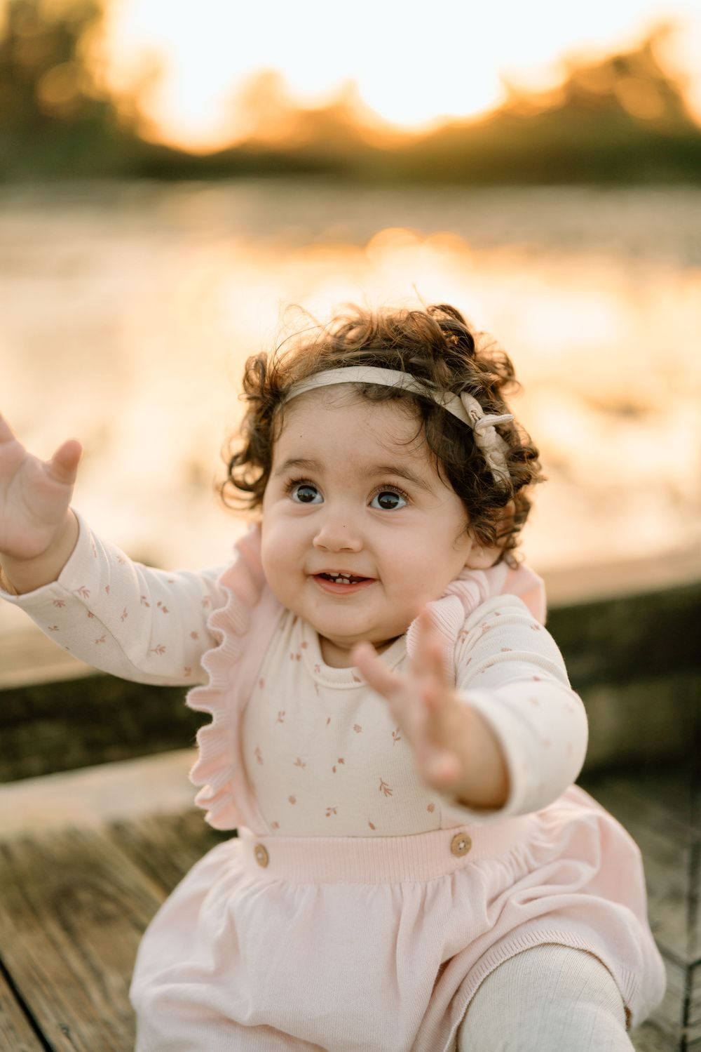 A baby in a pink dress and headband smiles joyfully while reaching out with both hands during golden hour sunset.