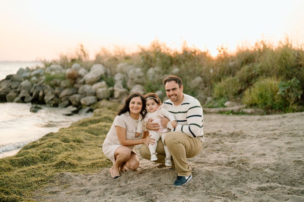 A family sits together on a beach at sunset with rocky outcroppings and grassy ground in the background.