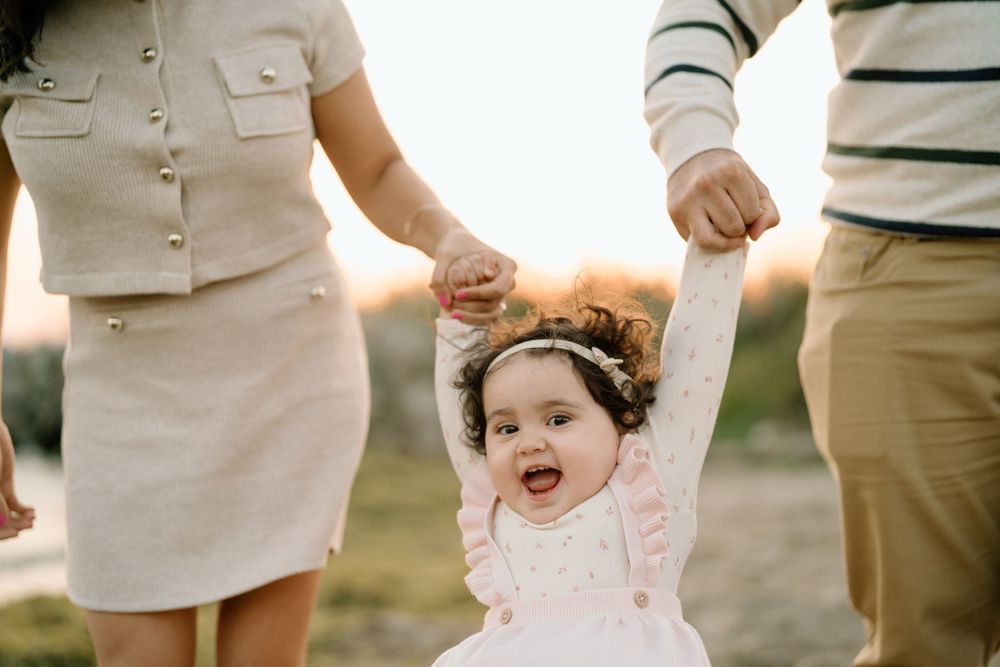 A small child wearing a pink dress laughs joyfully while being held by their parents' hands during golden hour.