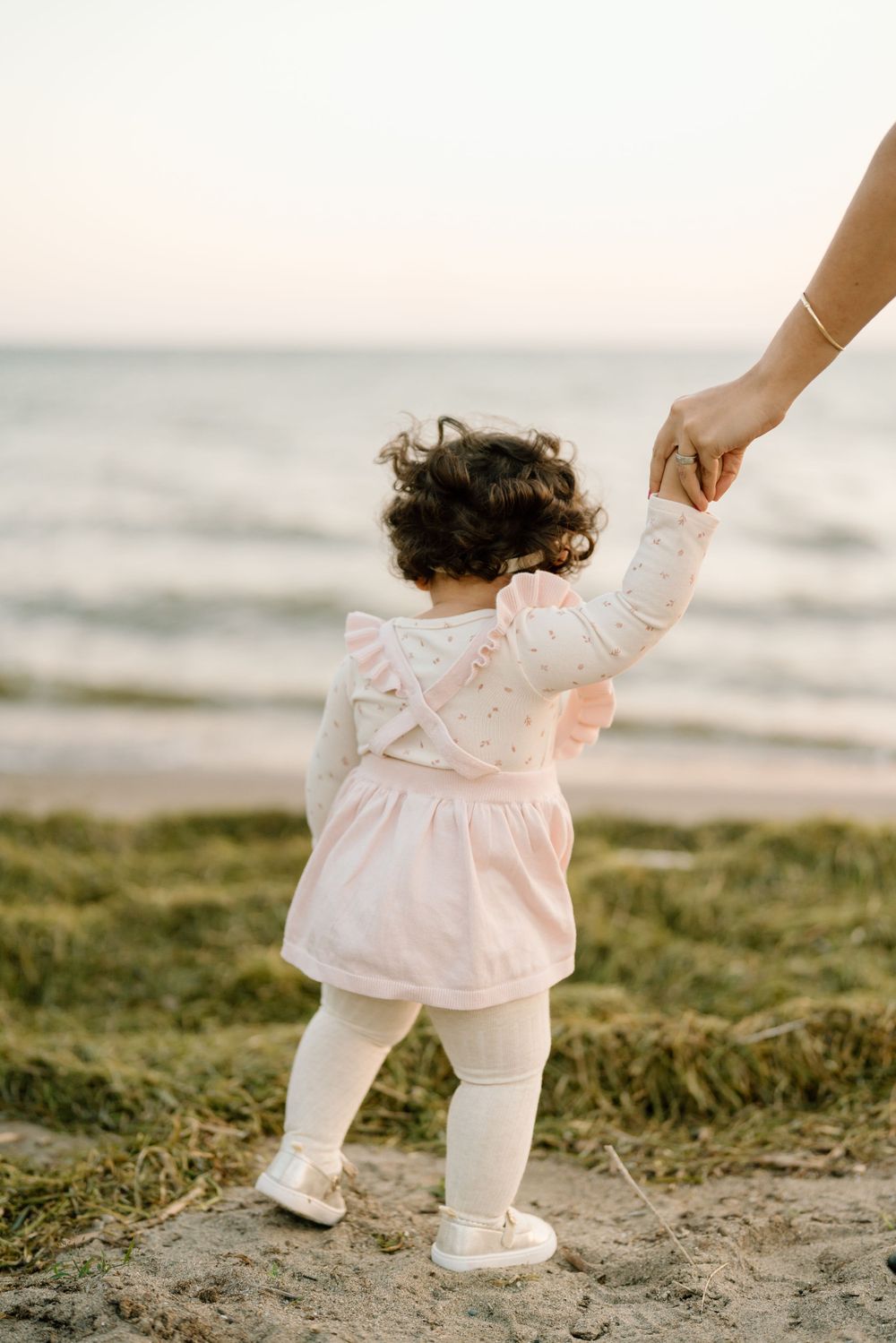 Small figure in pink dress and white tights holds an adult's hand while standing at the beach during sunset.