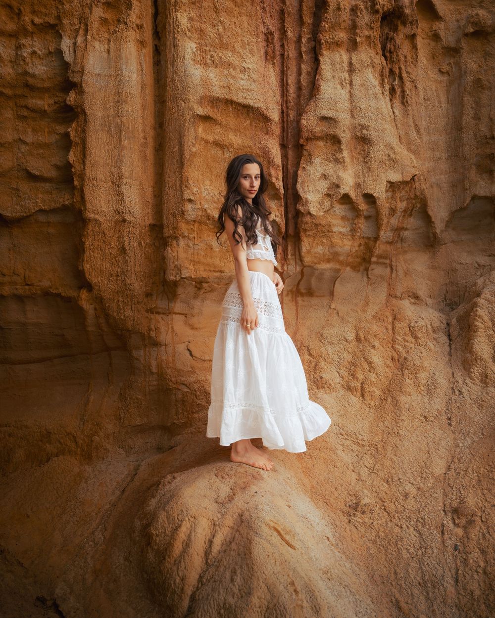 Figure in flowing white dress stands on rock formation in desert landscape.