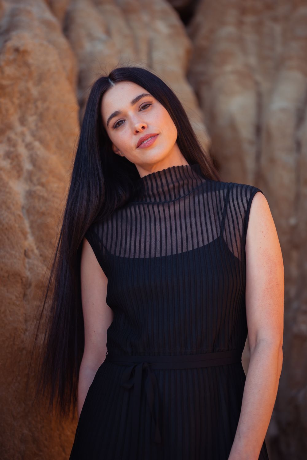 Portrait against textured rock wall wearing black ribbed high neck top in warm lighting.