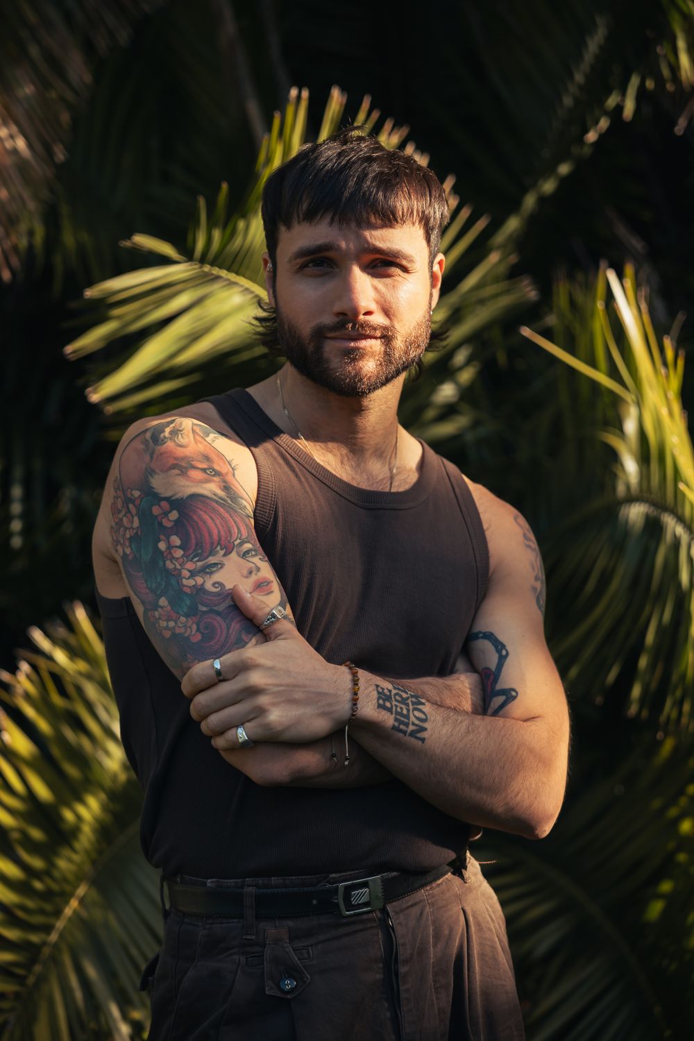 Person with colorful arm tattoos wearing dark tank top against tropical foliage.