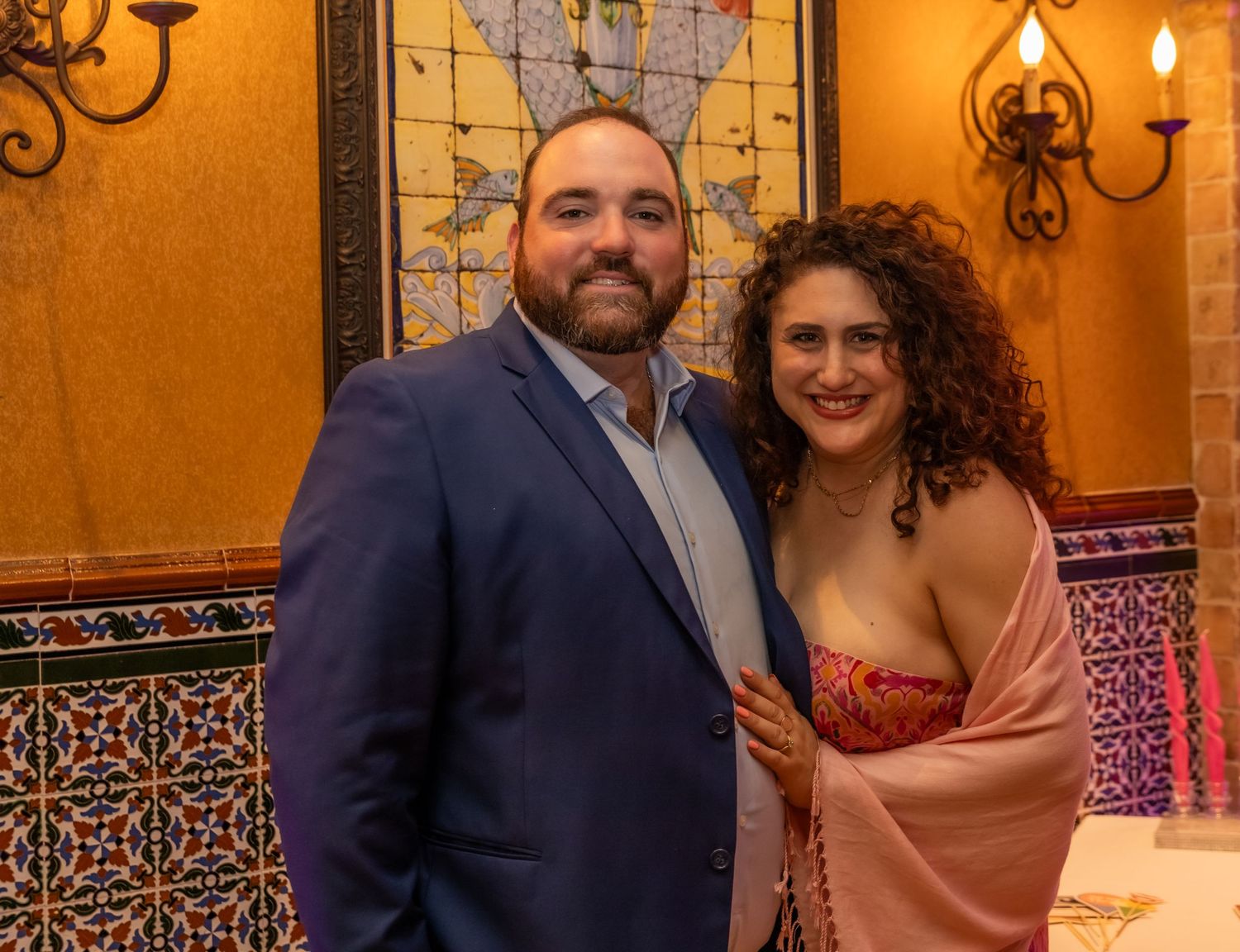A couple poses together against an orange wall with decorative wall sconces and ornate tile work in a warm-lit setting.